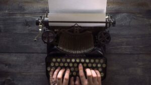 Overhead view of a vintage Corona typewriter with a blank sheet of paper loaded, hands typing on the keys.