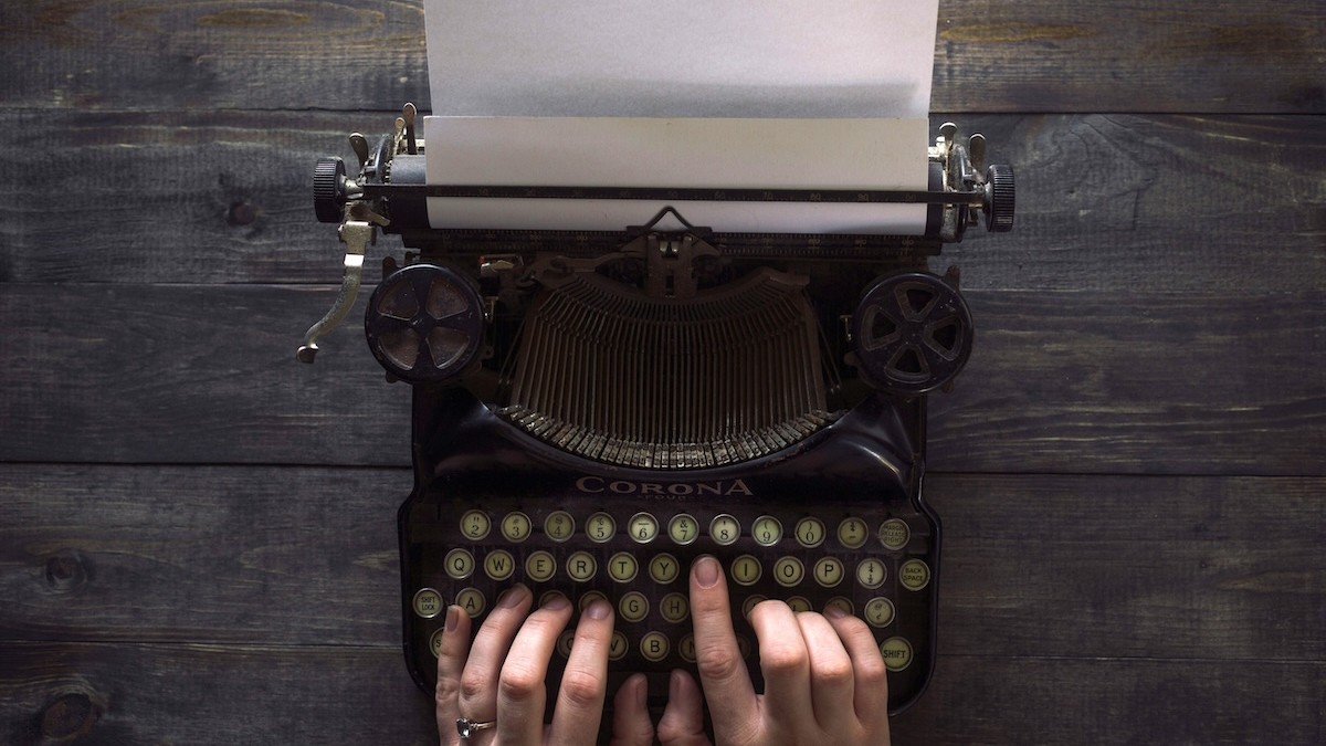Overhead view of a vintage Corona typewriter with a blank sheet of paper loaded, hands typing on the keys.