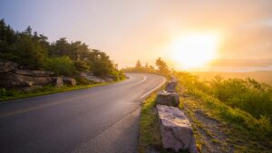 A curving two-lane road along a hillside at sunrise, with pine trees and granite boulders beside it and a hazy golden sun over a bay.